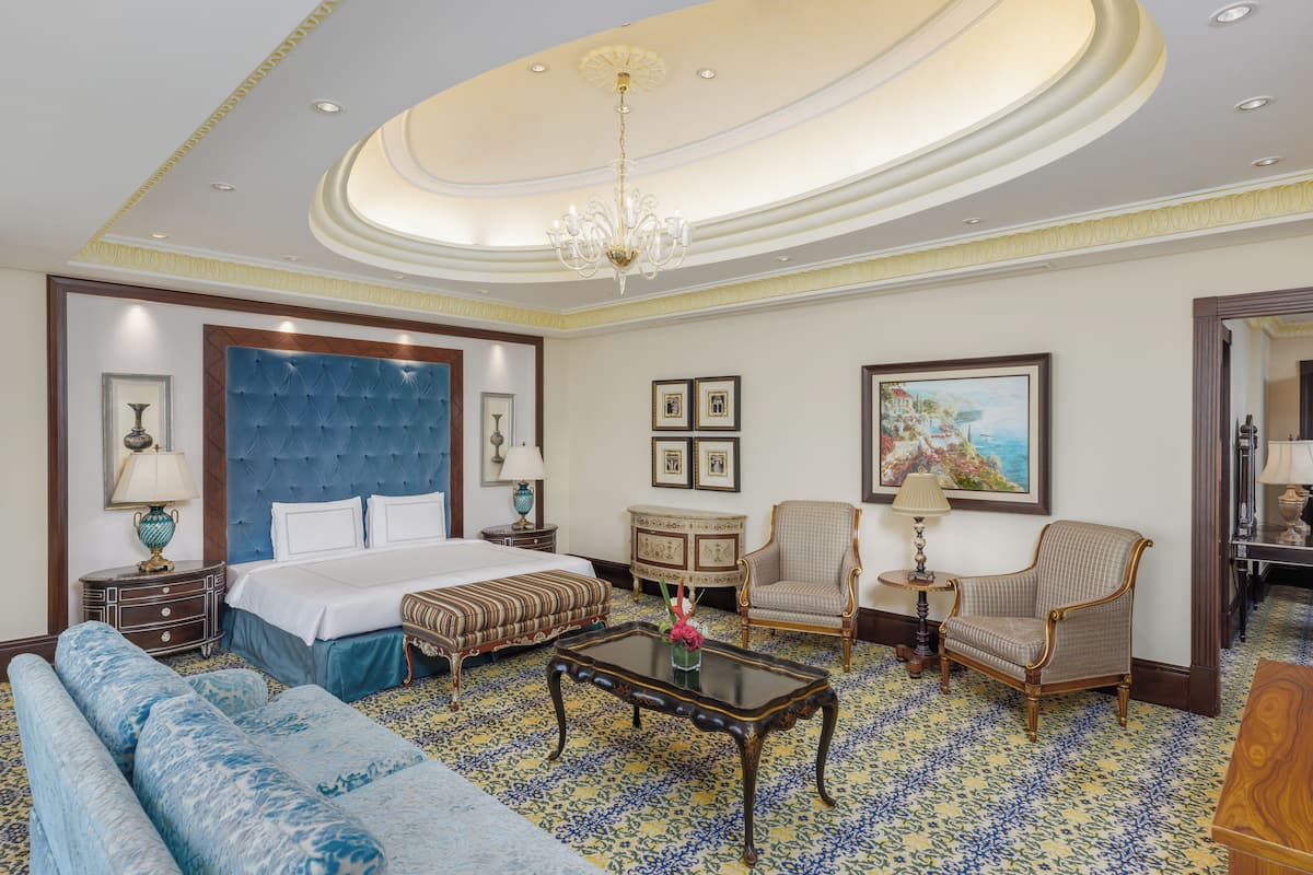 A hotel room with a large bed, a sofa, a coffee table, two chairs, a small table with a lamp, and a painting on the wall.