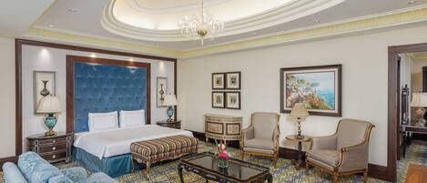 A hotel room with a large bed, a sofa, a coffee table, two chairs, a small table with a lamp, and a painting on the wall.