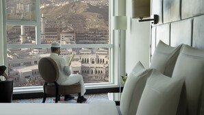 Un hombre sentado junto a un amplio ventanal en una habitación de hotel, con vistas a un paisaje urbano lleno de edificios históricos.