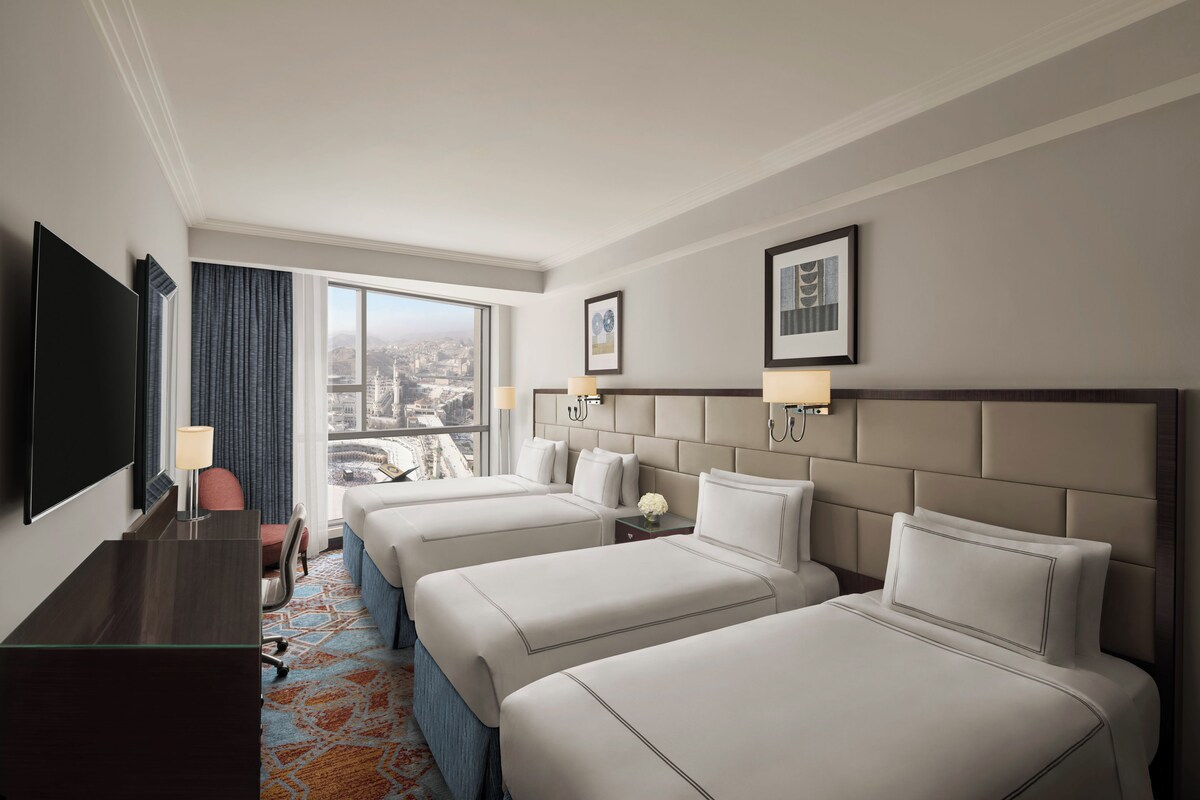 A hotel room with three beds, a desk, a TV, and a large window with a city view.
