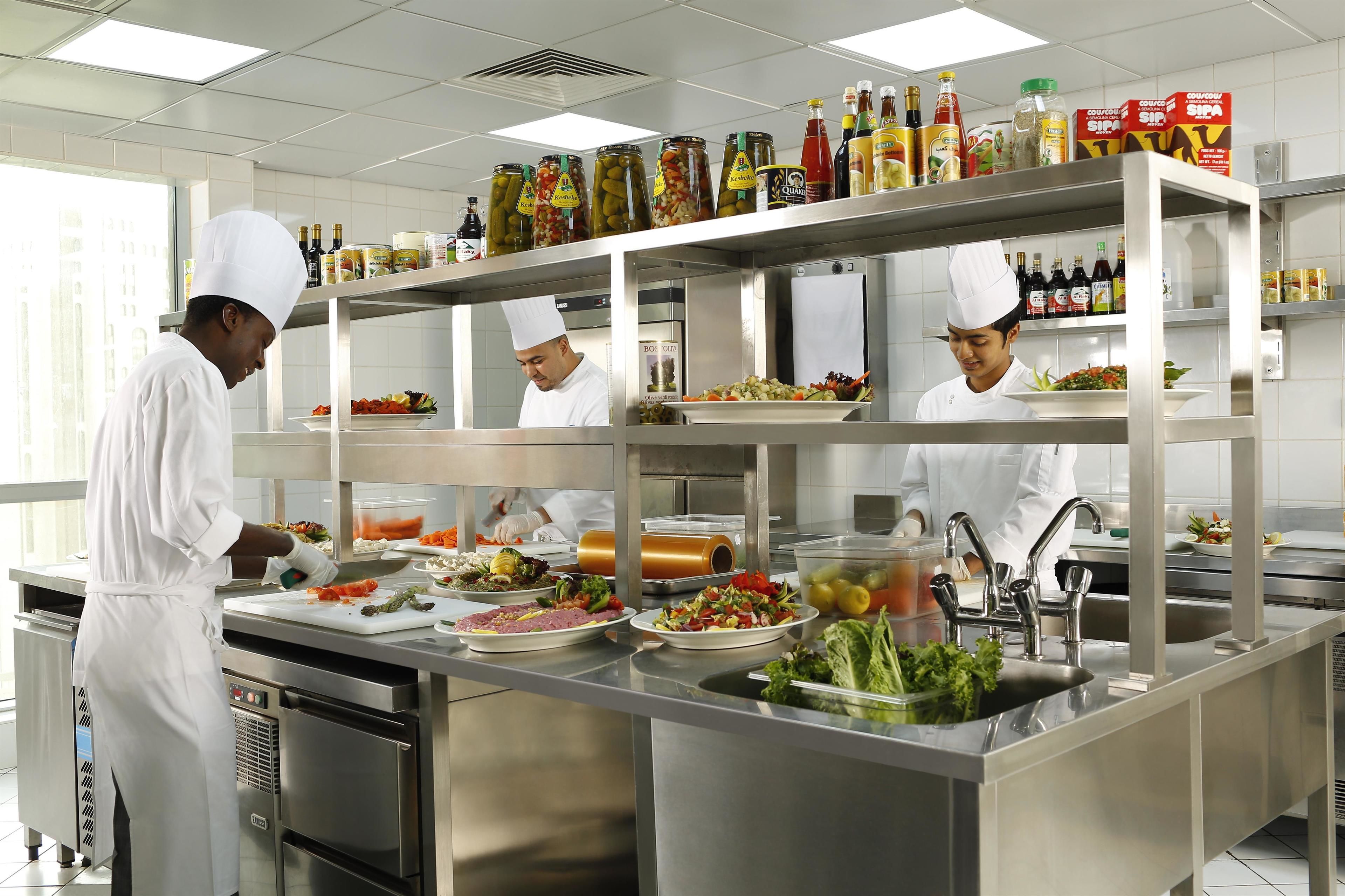 A professional kitchen with chefs preparing food, stainless steel appliances, and shelves stocked with condiments.
