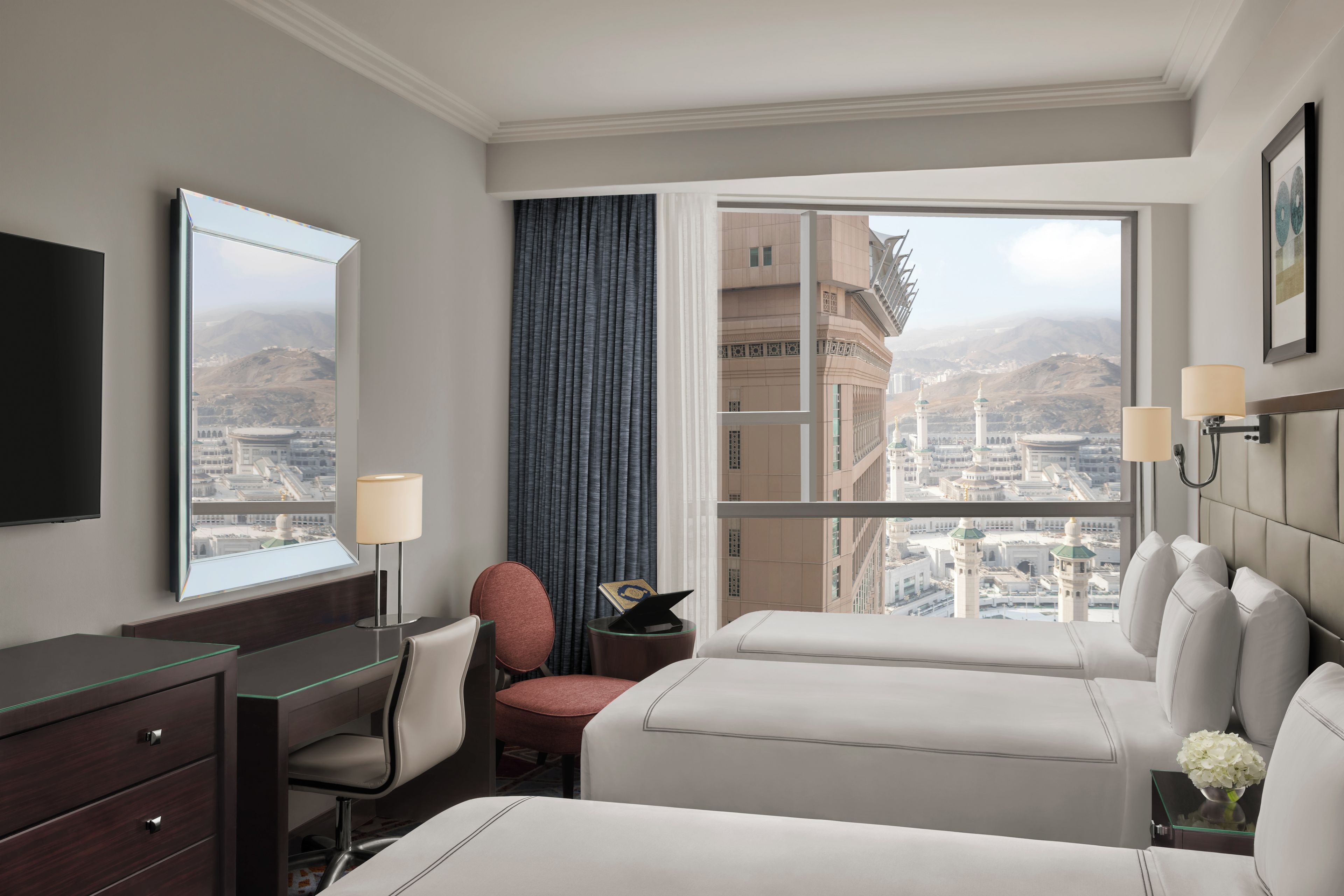 Una habitación de hotel con un ventanal grande, un escritorio, dos camas, una silla y una mesita de noche.