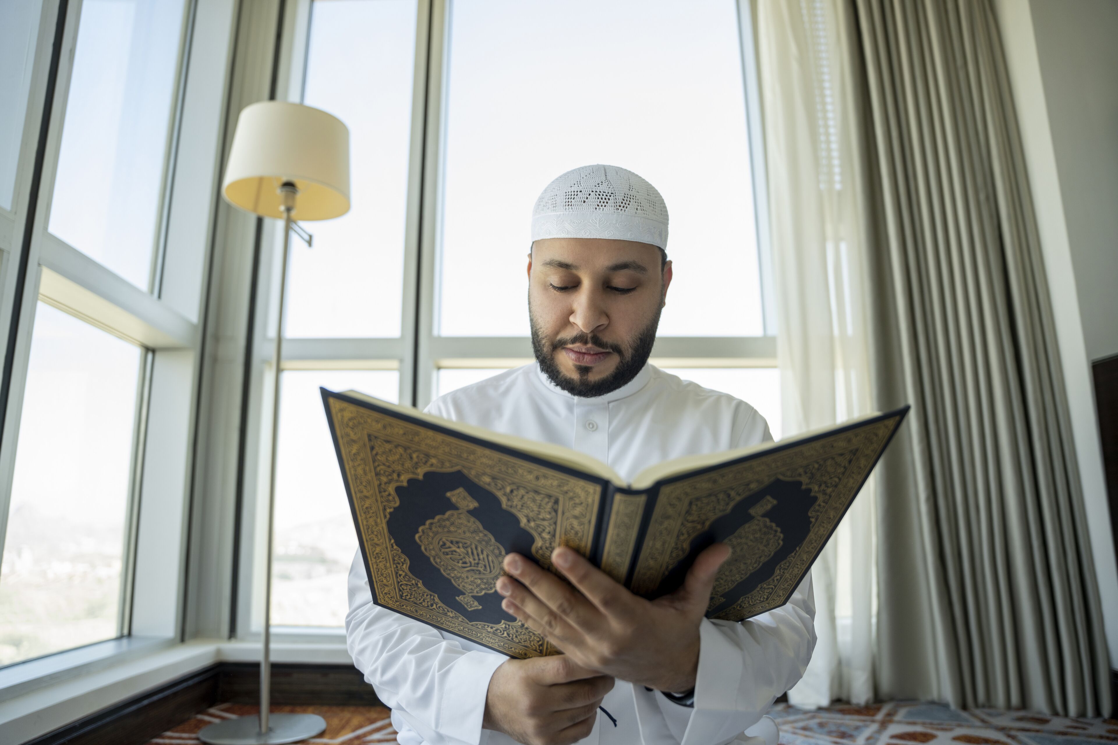 Una persona legge un libro in una stanza con un'ampia finestra, una lampada da terra e tende.