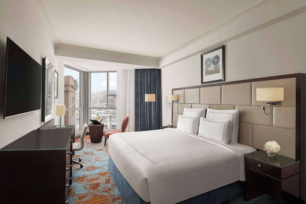 A hotel room with a large bed, a desk, a chair, a TV, and a view of the city.
