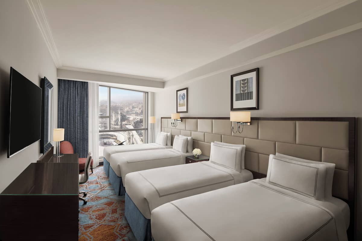 A hotel room with three beds, a desk, a TV, and a large window with a city view.