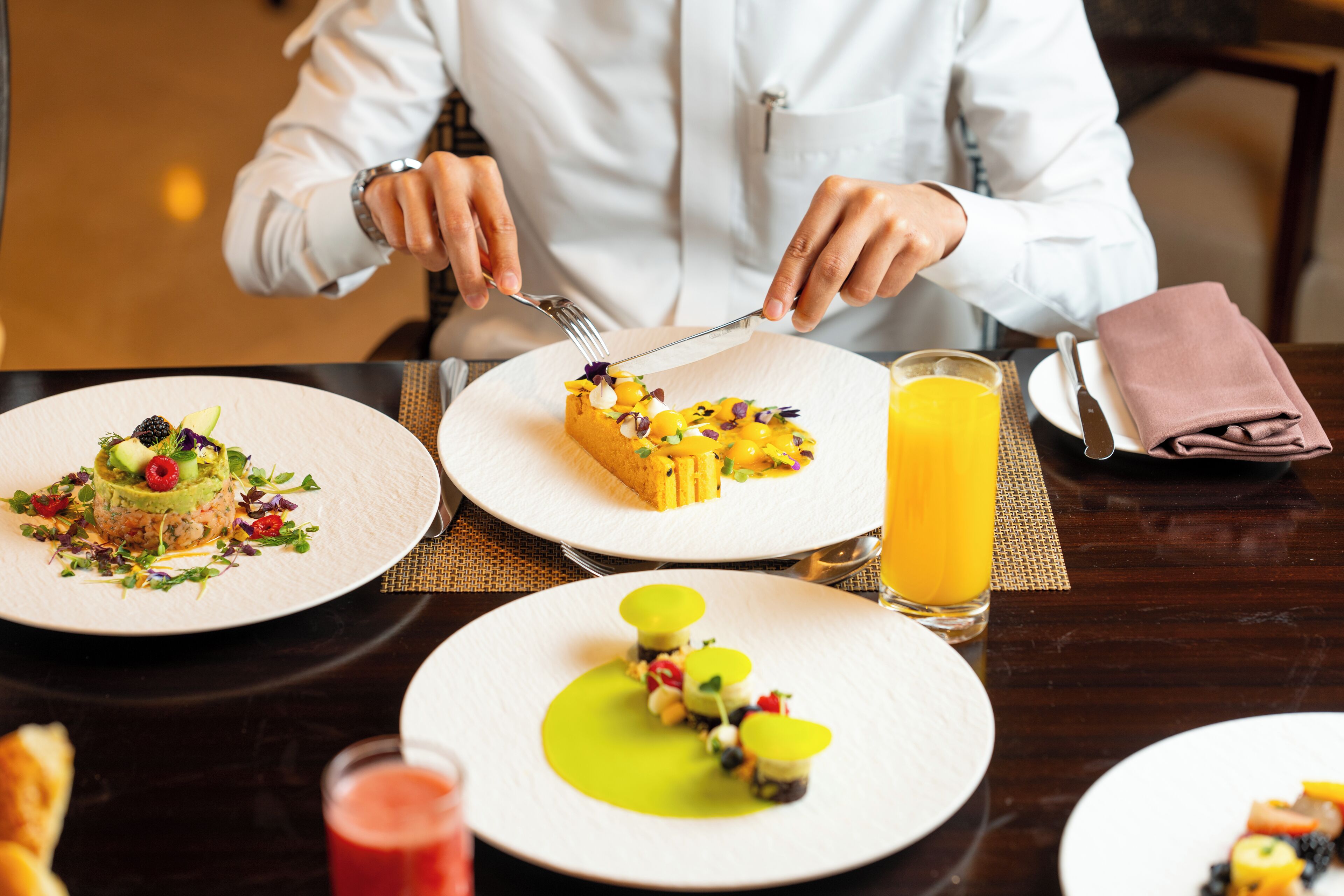 A person dining at a table with various dishes, including a dessert with fruit and a green sauce, and a glass of orange juice.