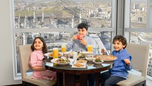 Una mesa de comedor con comida y bebidas, una vista de la ciudad a través de amplias ventanas y un paisaje urbano con edificios y una mezquita.