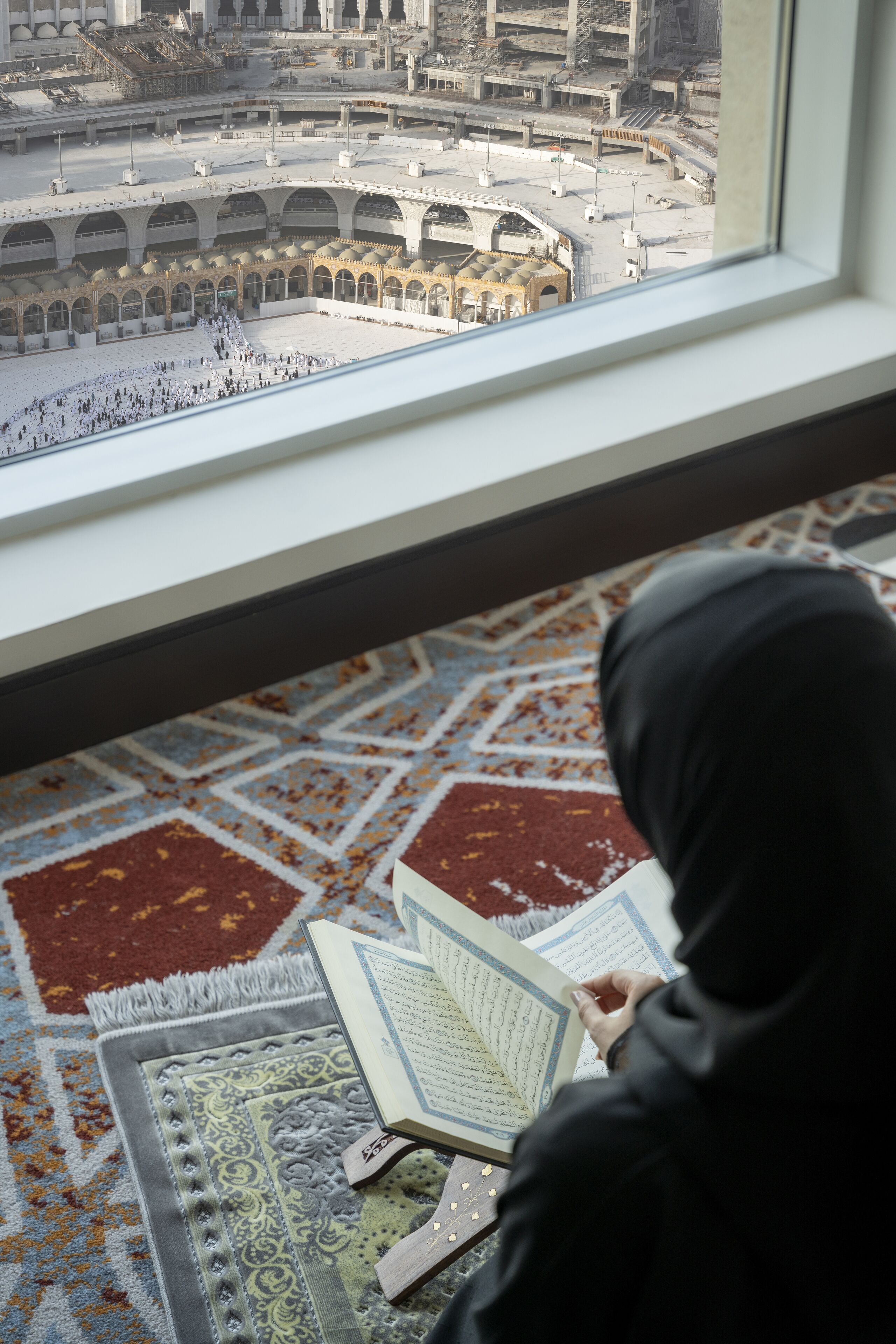 A view of a mosque's courtyard through a window, with a person reading a book.