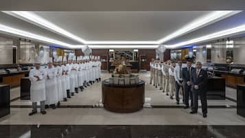 A formal dining area with a long table of waitstaff in white uniforms, a central decorative piece, and a modern chandelier.