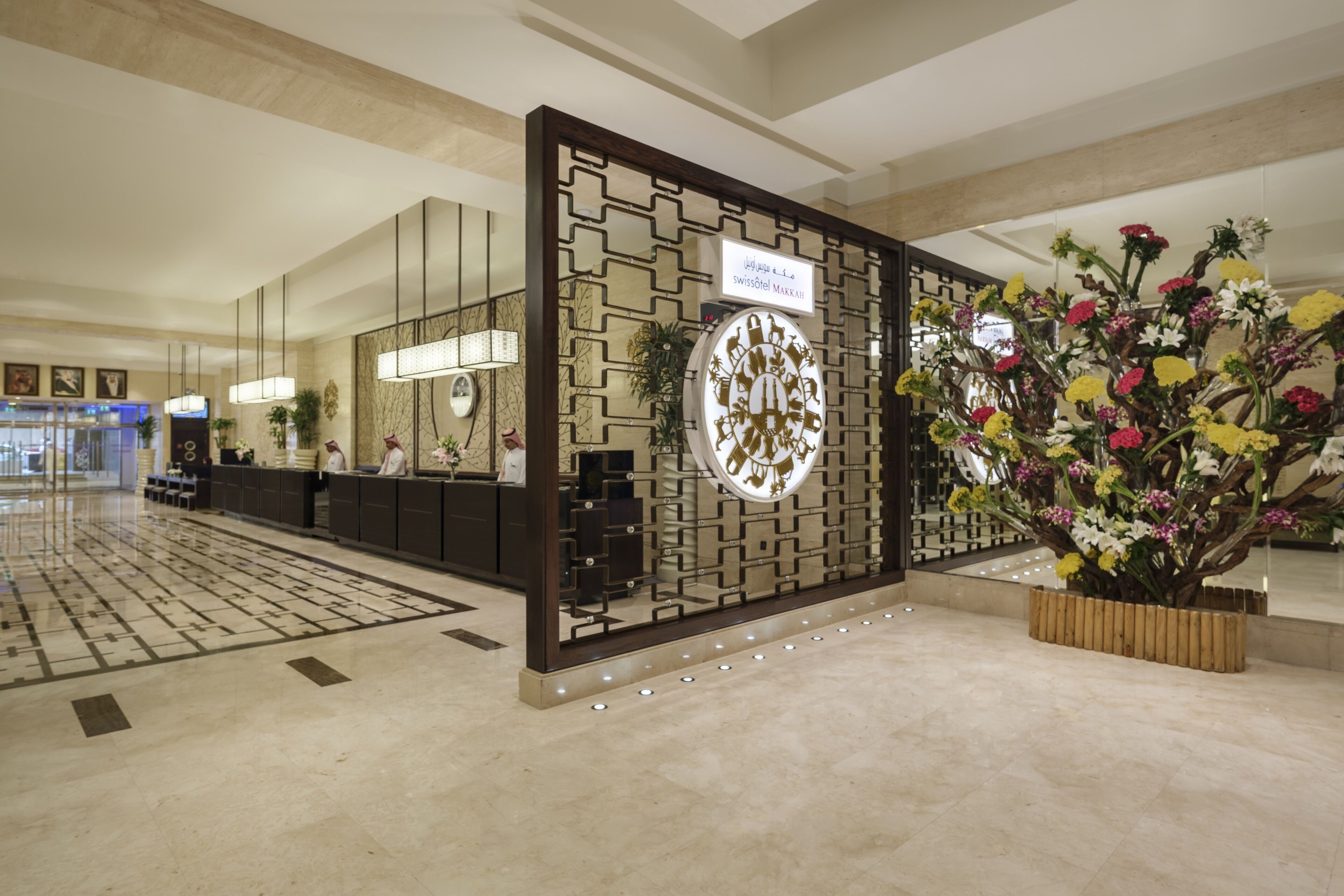 Un espace d’accueil d’hôtel au design moderne, comprenant un comptoir d’accueil, un mur décoratif agrémenté d’une horloge et un grand arrangement floral.