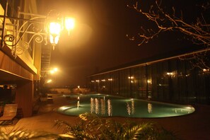 Un área de piscina iluminada de noche, con un muelle de madera, sillones de descanso y un edificio de vidrio al fondo.