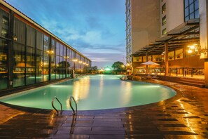 Piscina com cobertura de vidro, rodeada por um pátio de tijolos e edifícios vizinhos.