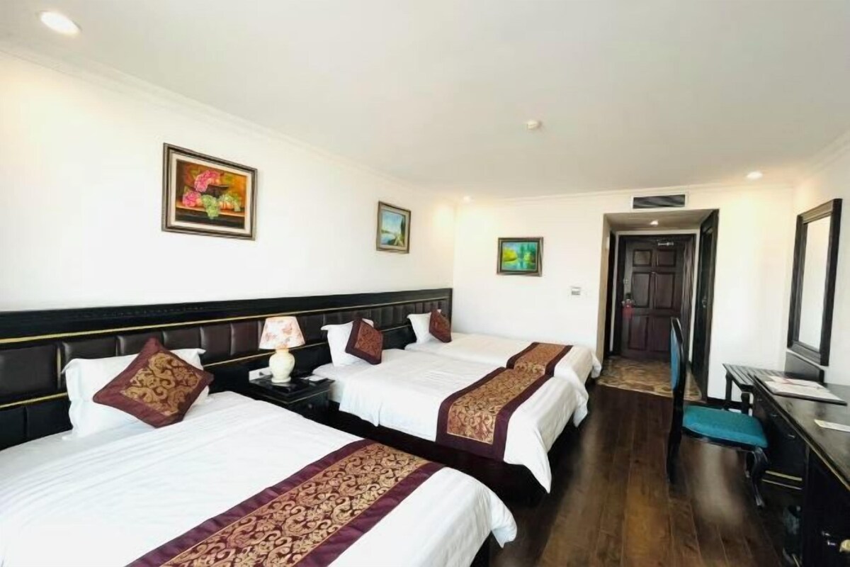 A hotel room with two beds, a desk, and a chair. There are paintings on the walls and a door in the background.