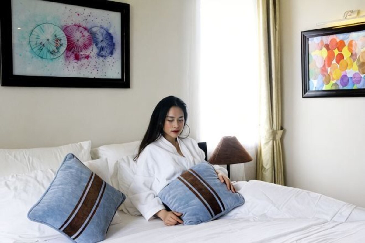 Una persona sentada en una cama con dos almohadas, una lámpara y dos cuadros en la pared.