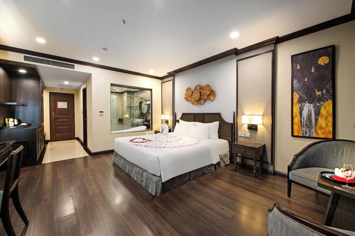 Een hotelkamer met een groot bed, een bureau, een stoel, een eettafel met taart en een schilderij aan de muur.