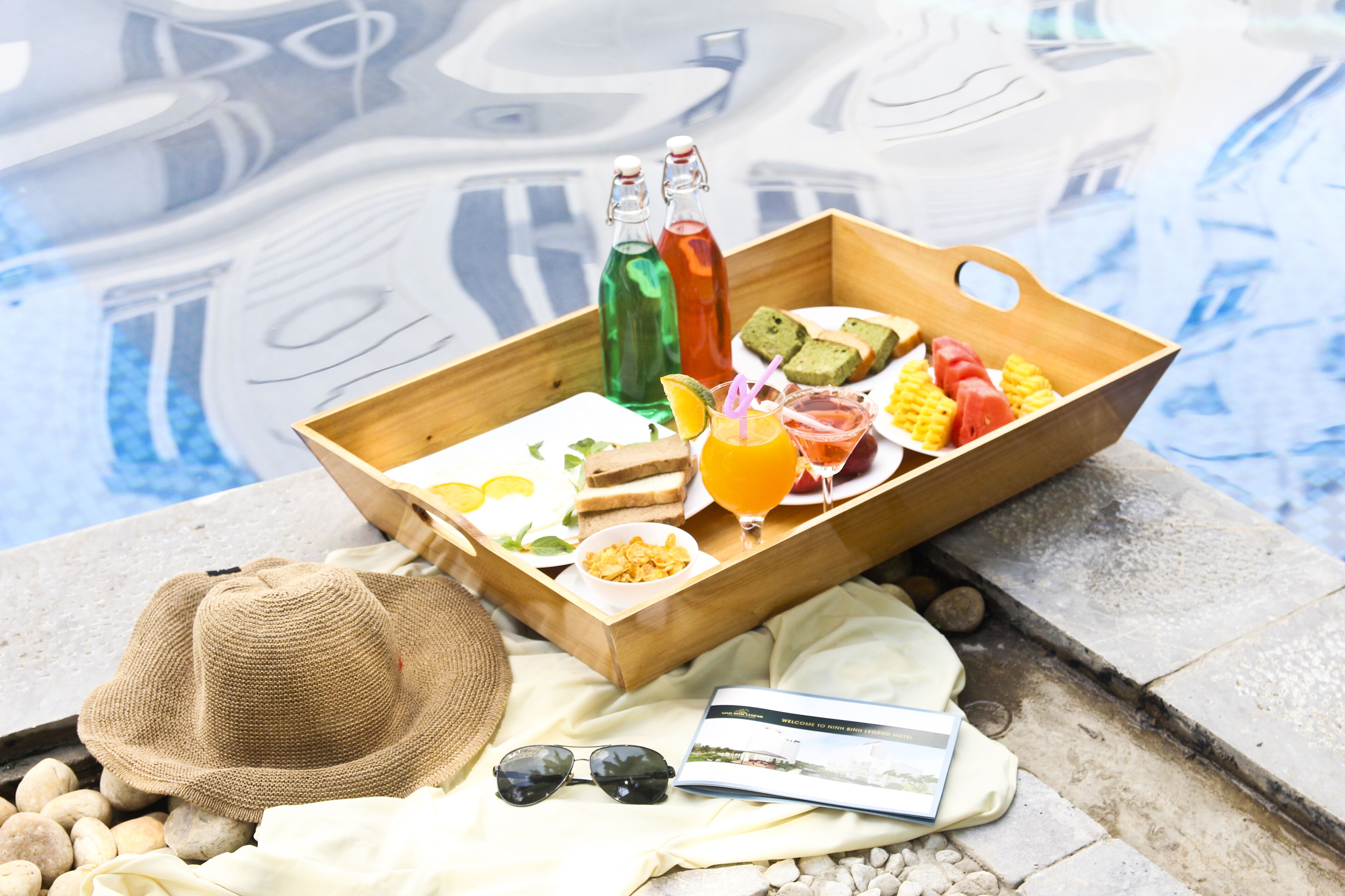 A wooden tray with food and drinks by a pool.