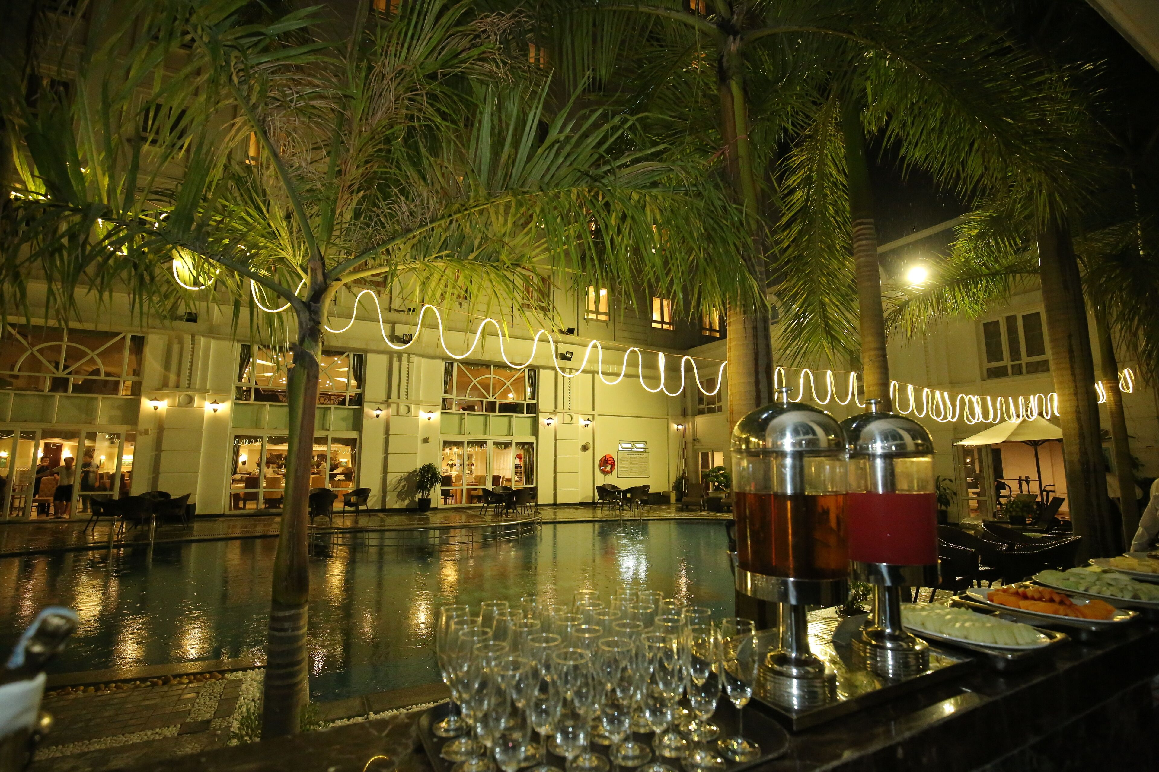 A poolside bar area with a buffet setup, palm trees, and a building with illuminated signage in the background.