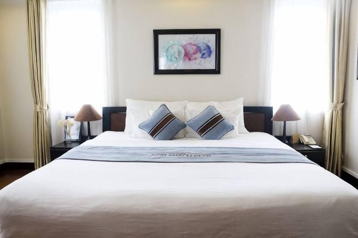 Een net opgemaakt bed met twee kussens, een lijst met een abstract schilderij erboven, en een nachtkastje met een lamp en een telefoon.