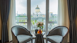 Une pièce comprenant deux fauteuils, une petite table avec un panier de fruits et des fleurs, et une vue sur un grand bâtiment coiffé d’un dôme, visible à travers une porte vitrée.
