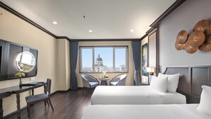 A hotel room with a large bed, a desk with a mirror, two chairs, a view of a cityscape, and decorative wall art.
