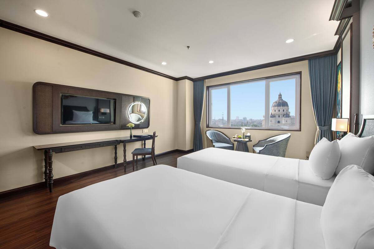 Een hotelkamer met twee bedden, een bureau met spiegel, een stoel, een televisie en een raam met uitzicht op de stad.