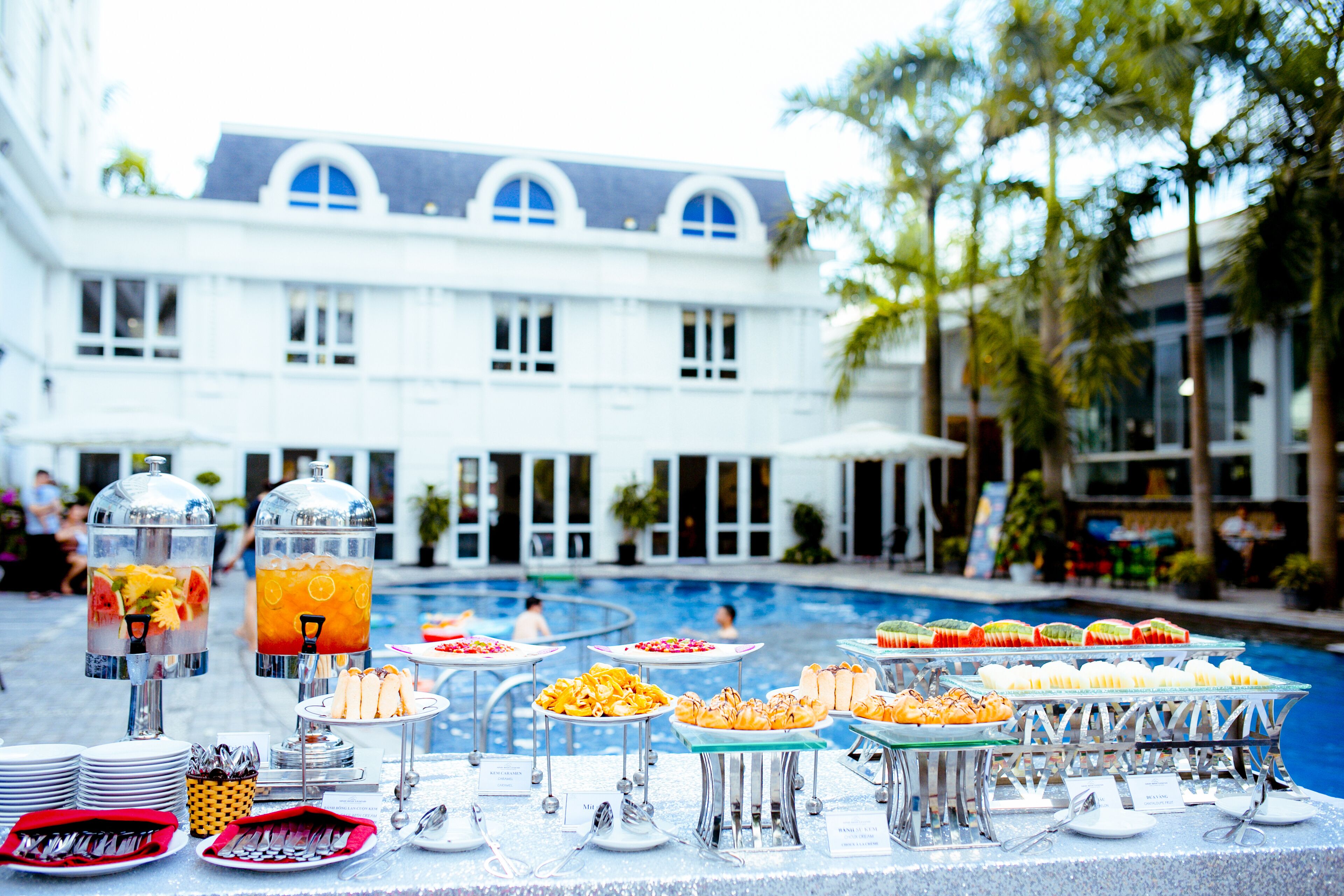A buffet setup with various food items and drinks, a pool, and a building in the background.