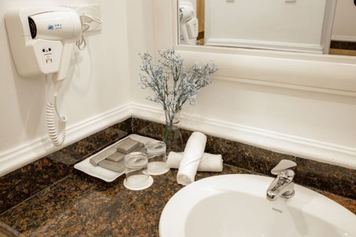 Un baño con un lavamanos blanco, un secador de pelo, un florero con flores secas y una toalla sobre la encimera.