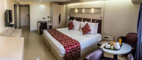 A hotel room with a bed, a desk with a chair, a TV, and a small table with a tray of breakfast items.