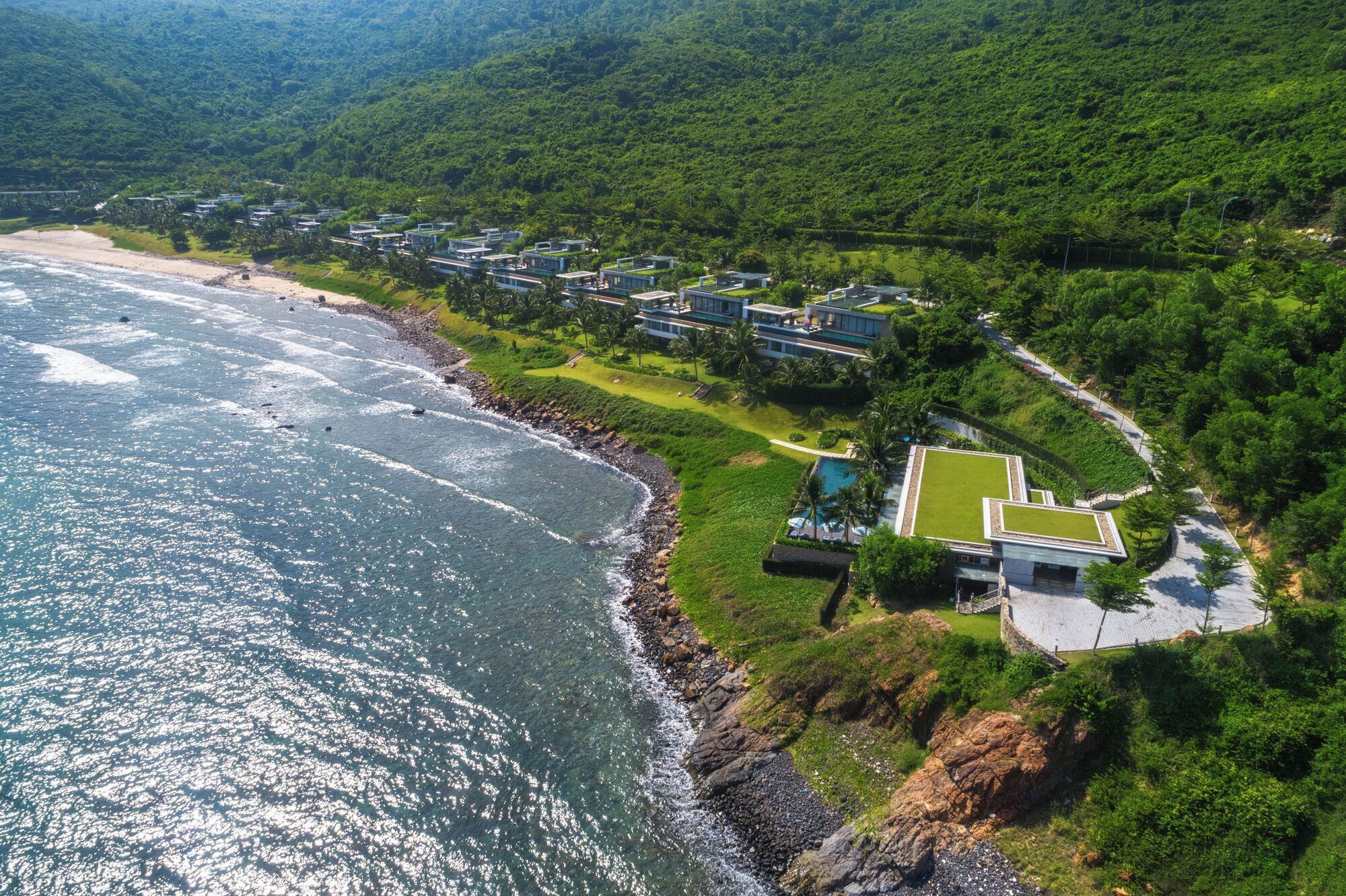 Vista aérea de un complejo turístico costero con villas modernas, una playa y abundante vegetación.