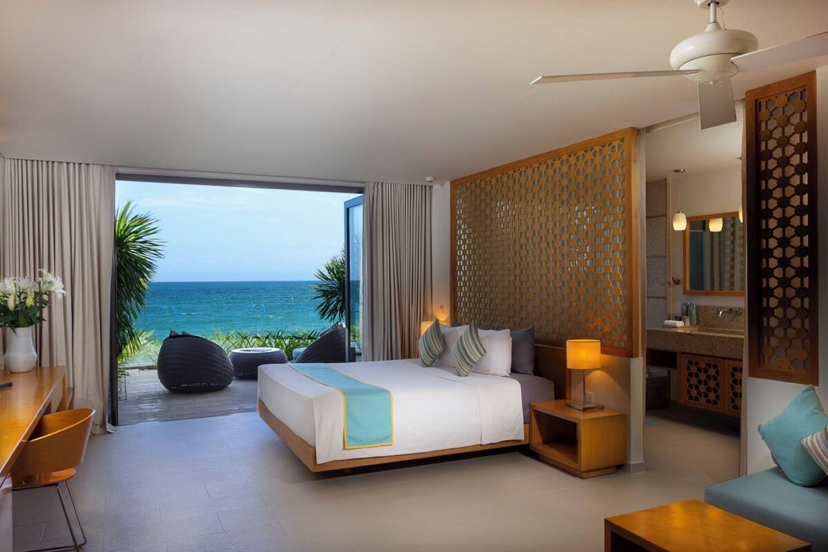 A hotel room with a bed, a desk, a chair, a sofa, a nightstand, a lamp, and a view of the ocean.