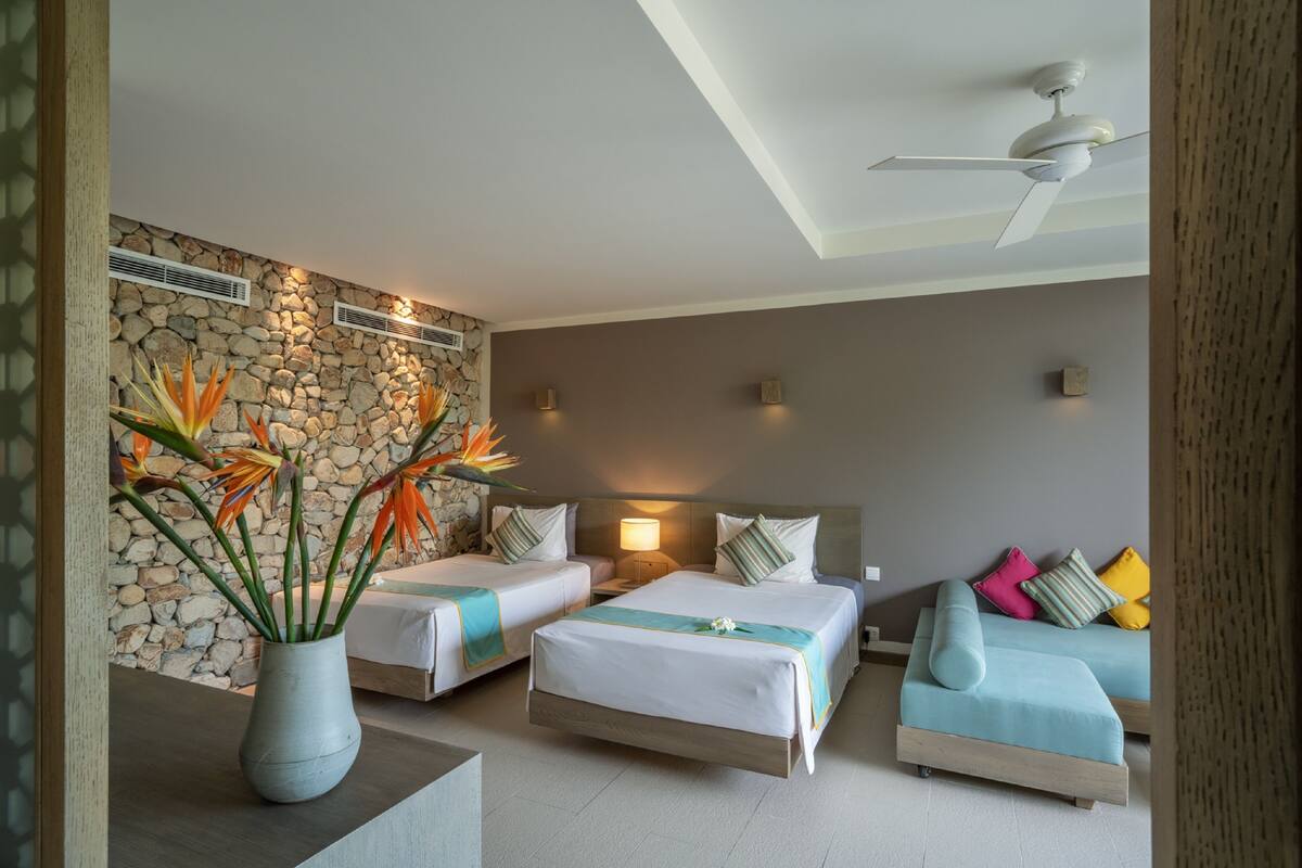 A hotel room with two beds, a sofa, a stone wall, and a ceiling fan.