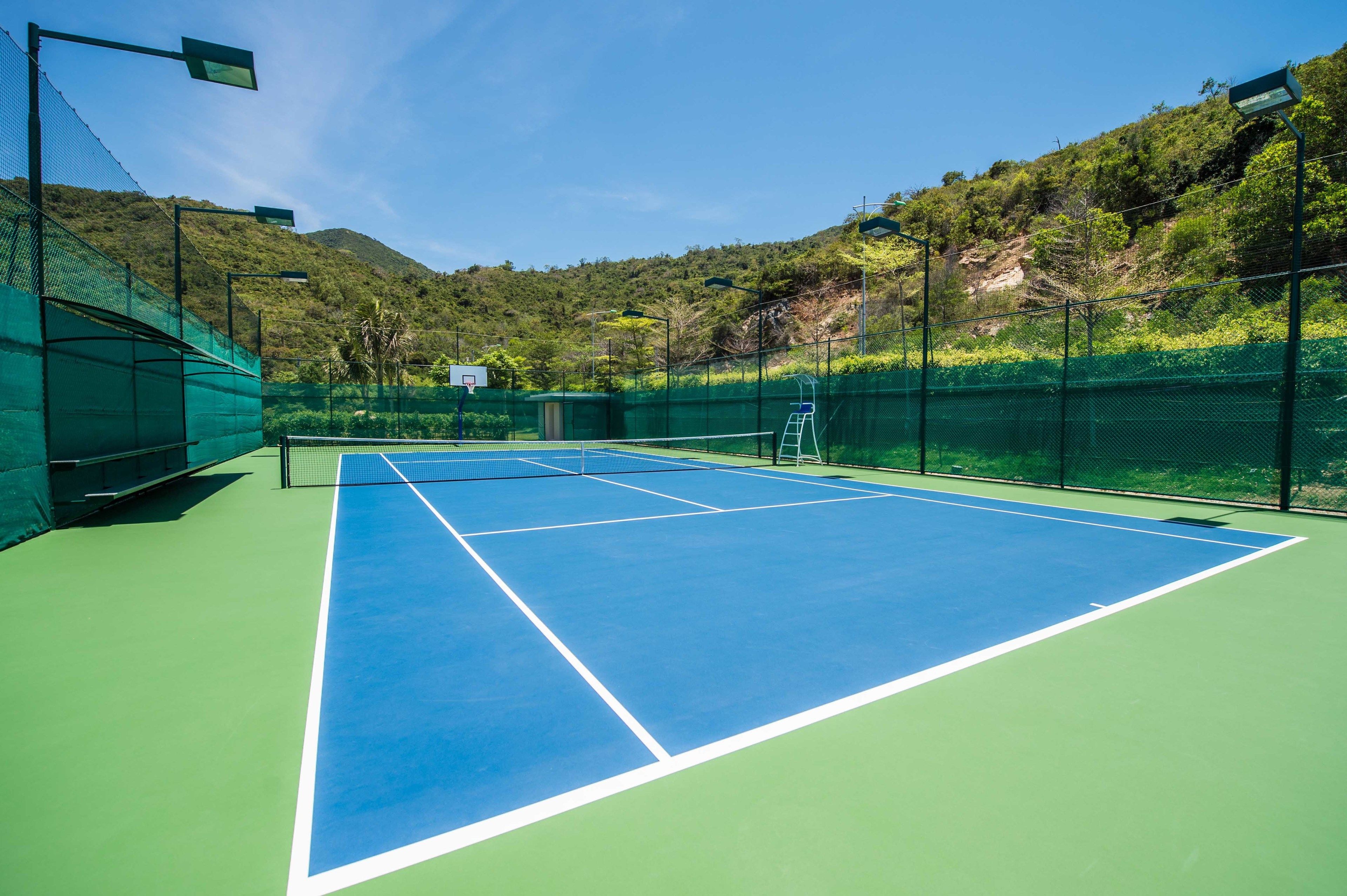 A tennis court with a net, surrounded by green fencing and a mountain backdrop.