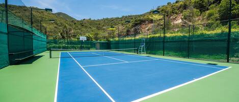 A tennis court with a net, surrounded by green fencing and a mountain backdrop.