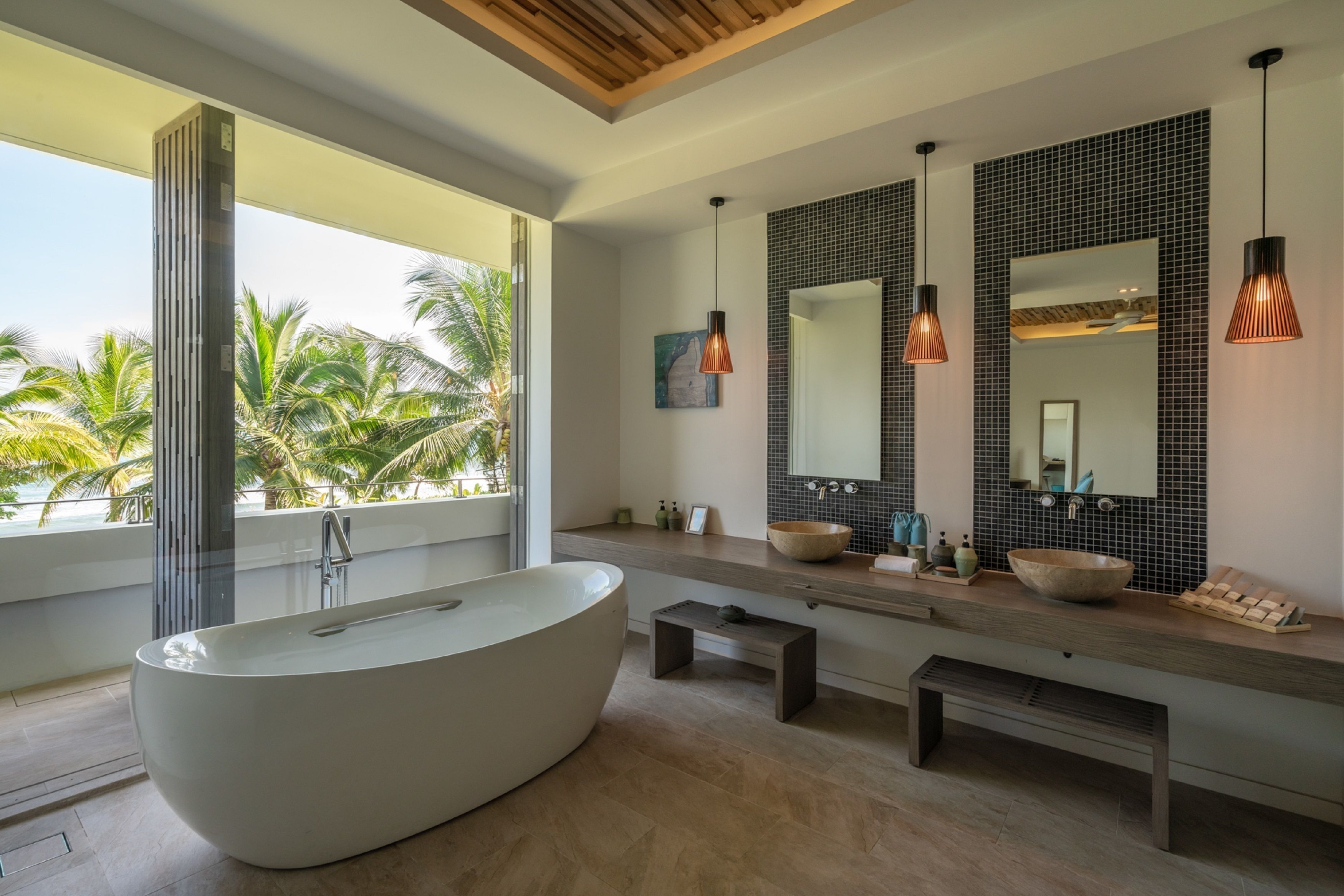 A modern bathroom with a freestanding bathtub, large mirrors, and a view of palm trees.