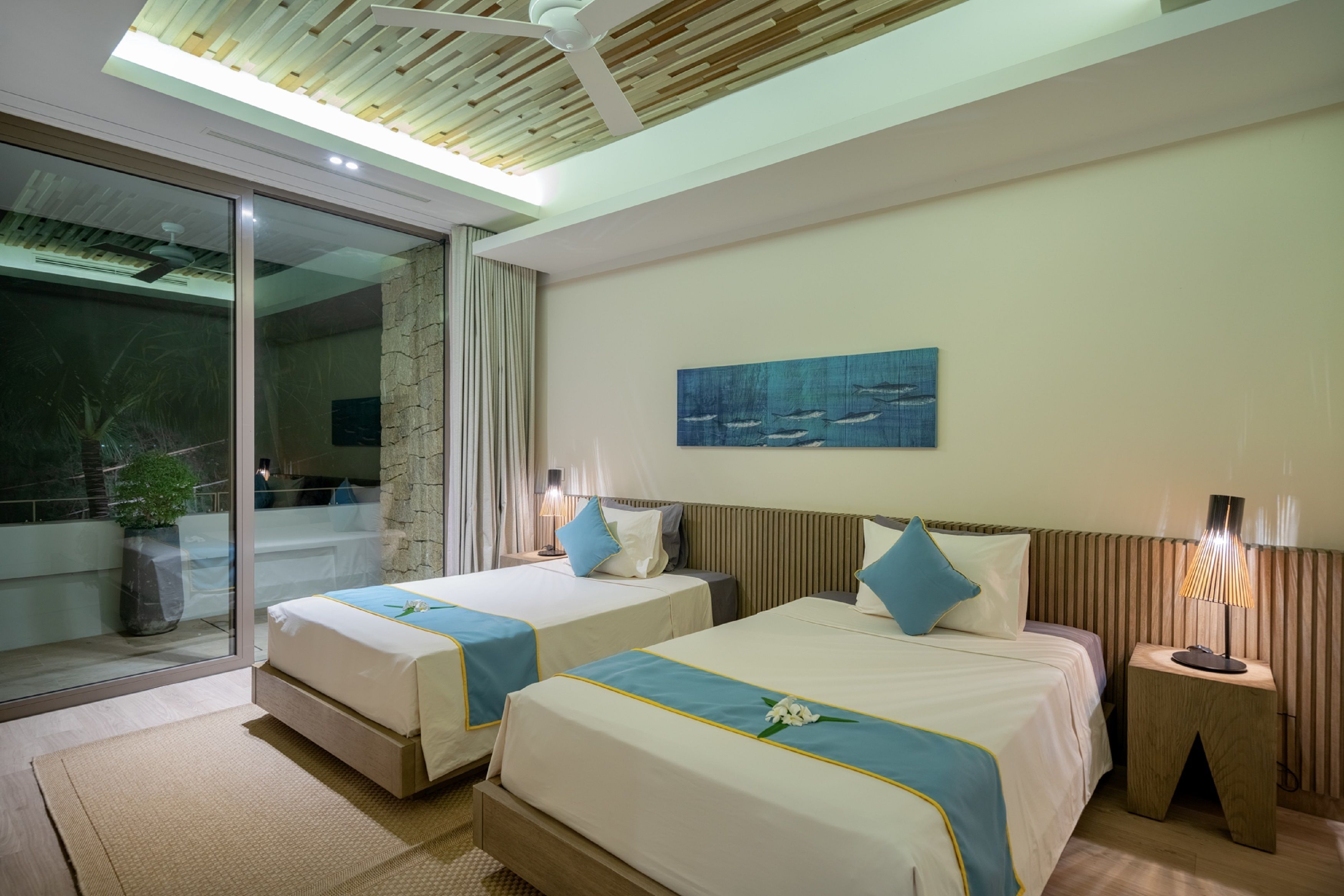 A hotel room with two beds, a large window, a ceiling fan, and a painting on the wall.