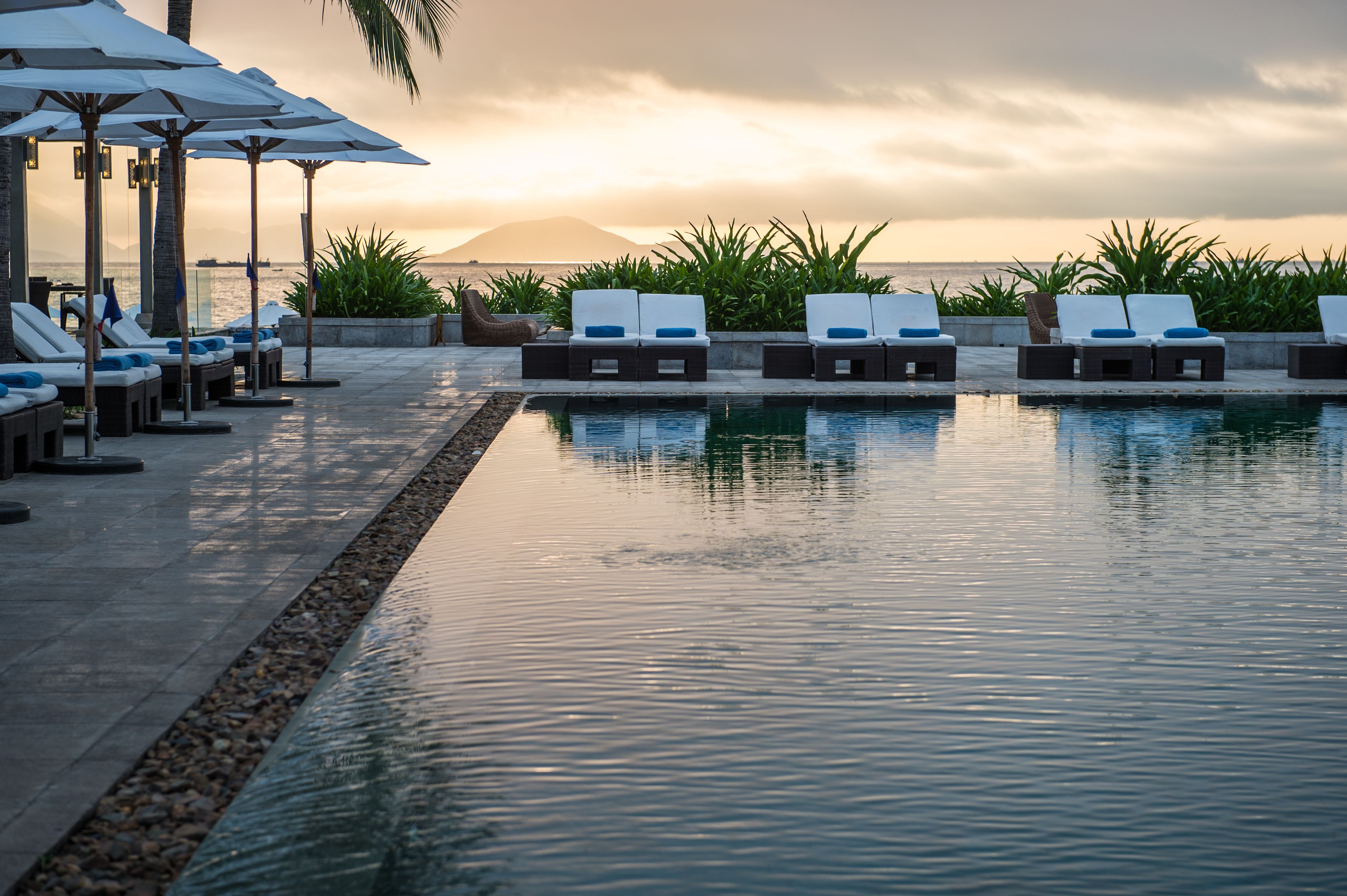 A poolside area with lounge chairs and umbrellas, a clear view of the ocean, and a sunset in the background.