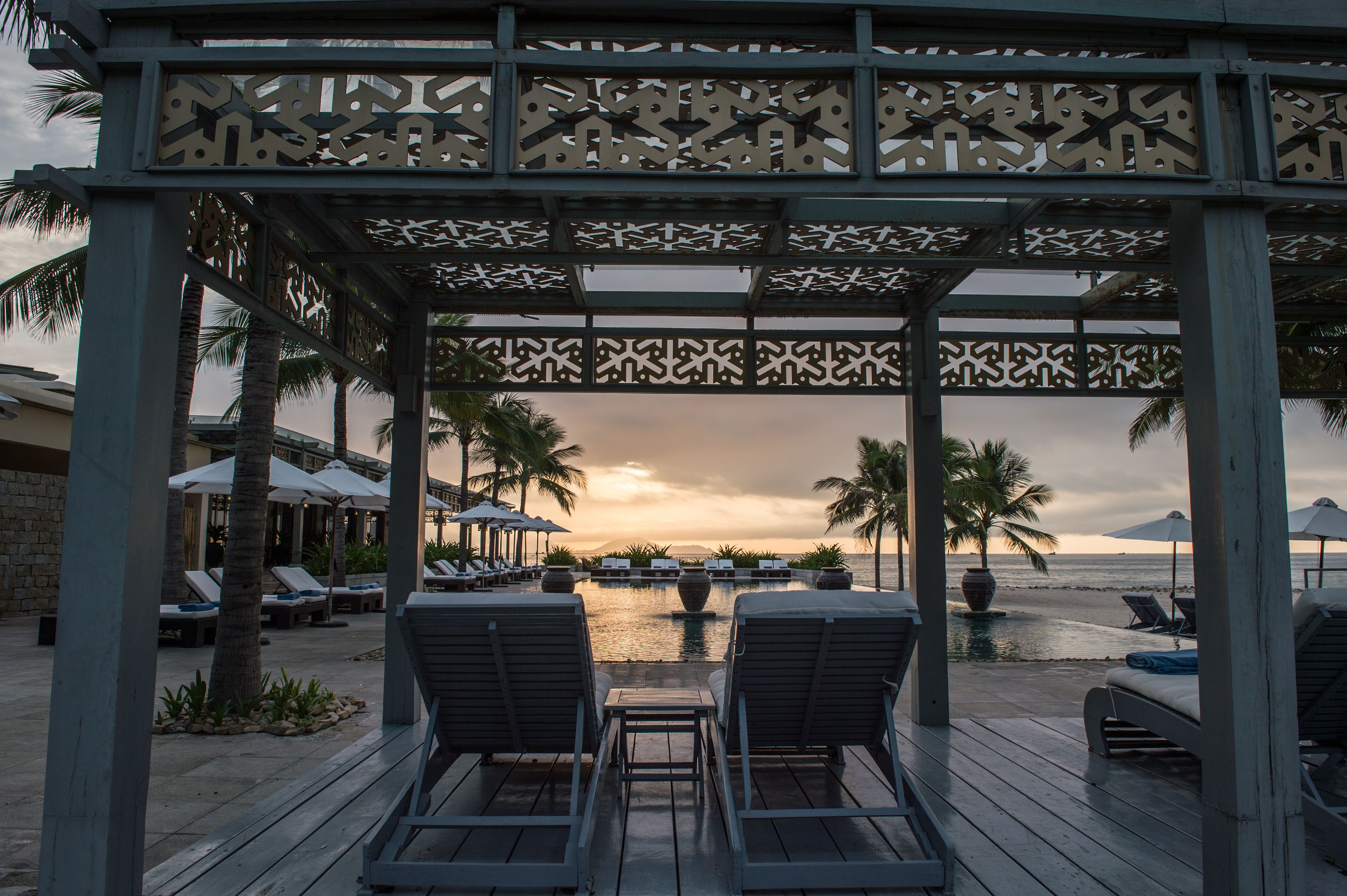 A wooden deck with lounge chairs and umbrellas overlooking a pool and beach, with palm trees and a sunset in the background.