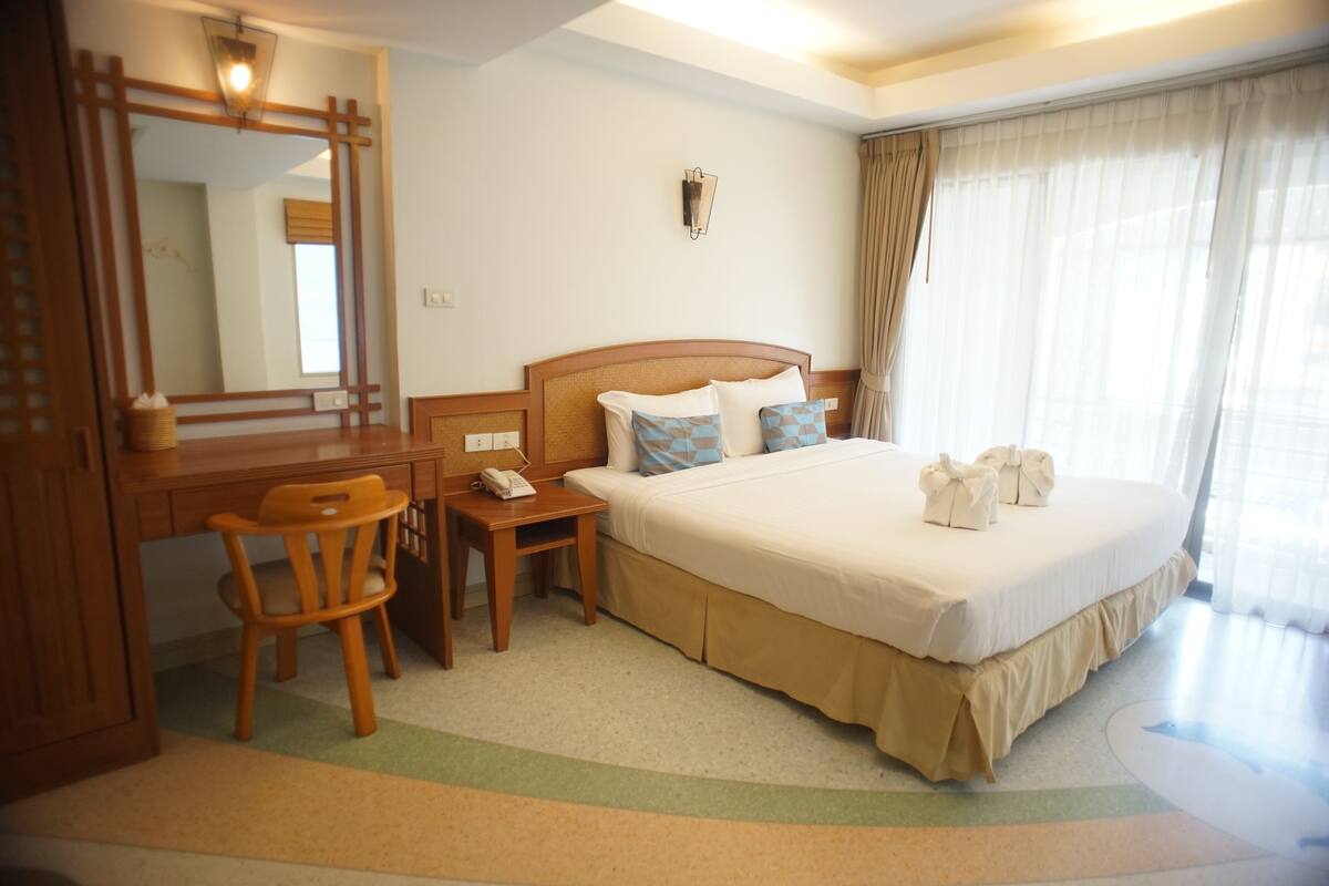 A hotel room with a large bed, a desk with a chair, a bedside table, a mirror, and a window with curtains.