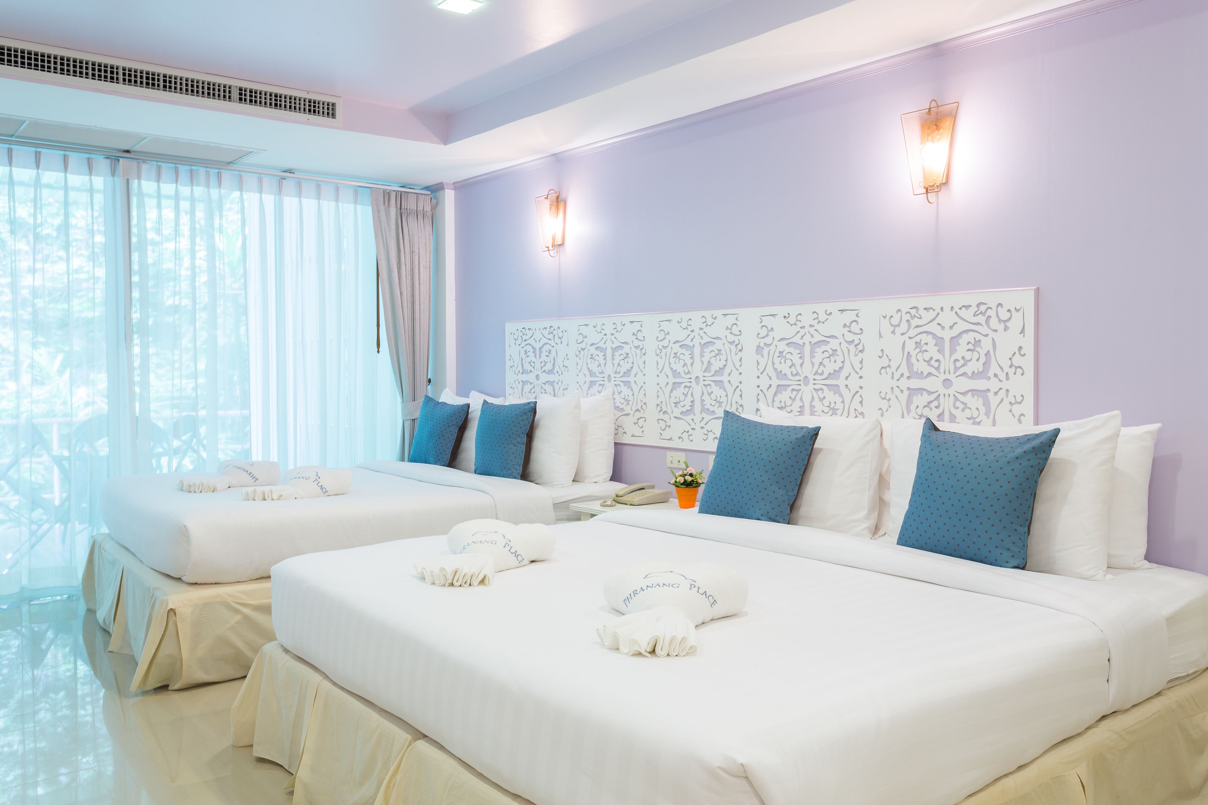 A hotel room with two beds, a large window with sheer curtains, a decorative headboard, and a wall-mounted light fixture.