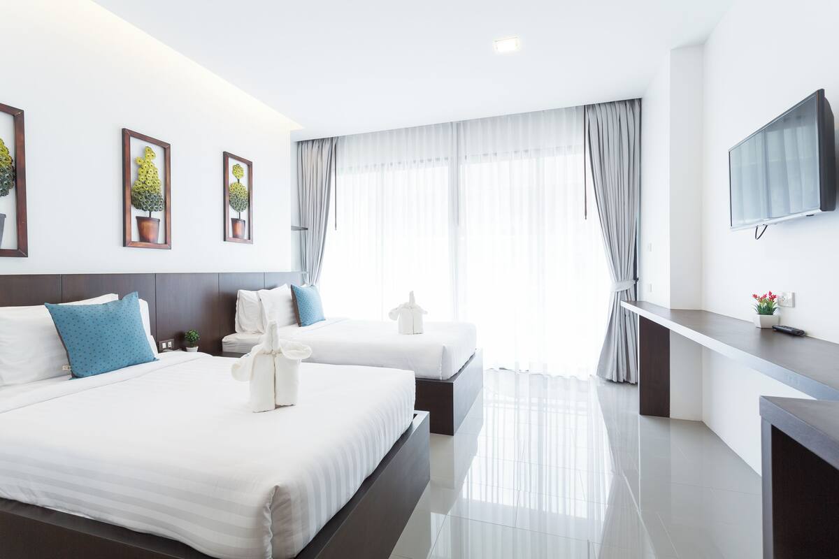 A modern hotel room with two beds, a flat-screen TV, a small table, and a large window with curtains.