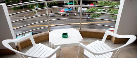 A balcony with two white plastic chairs and a small table, overlooking a busy street with vehicles and pedestrians.