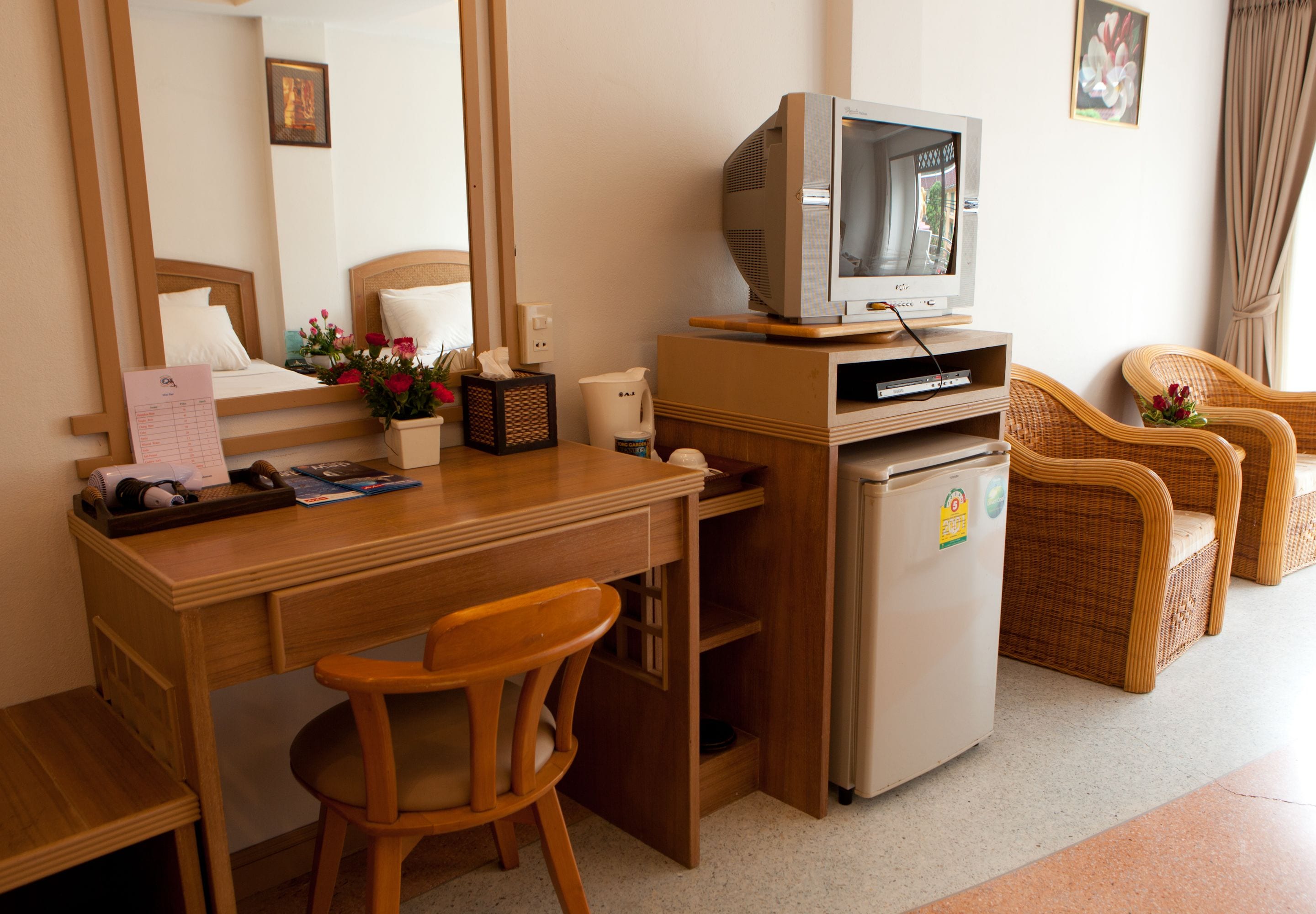 A hotel room with a wooden desk, a television, a refrigerator, wicker chairs, and a bed with white bedding.
