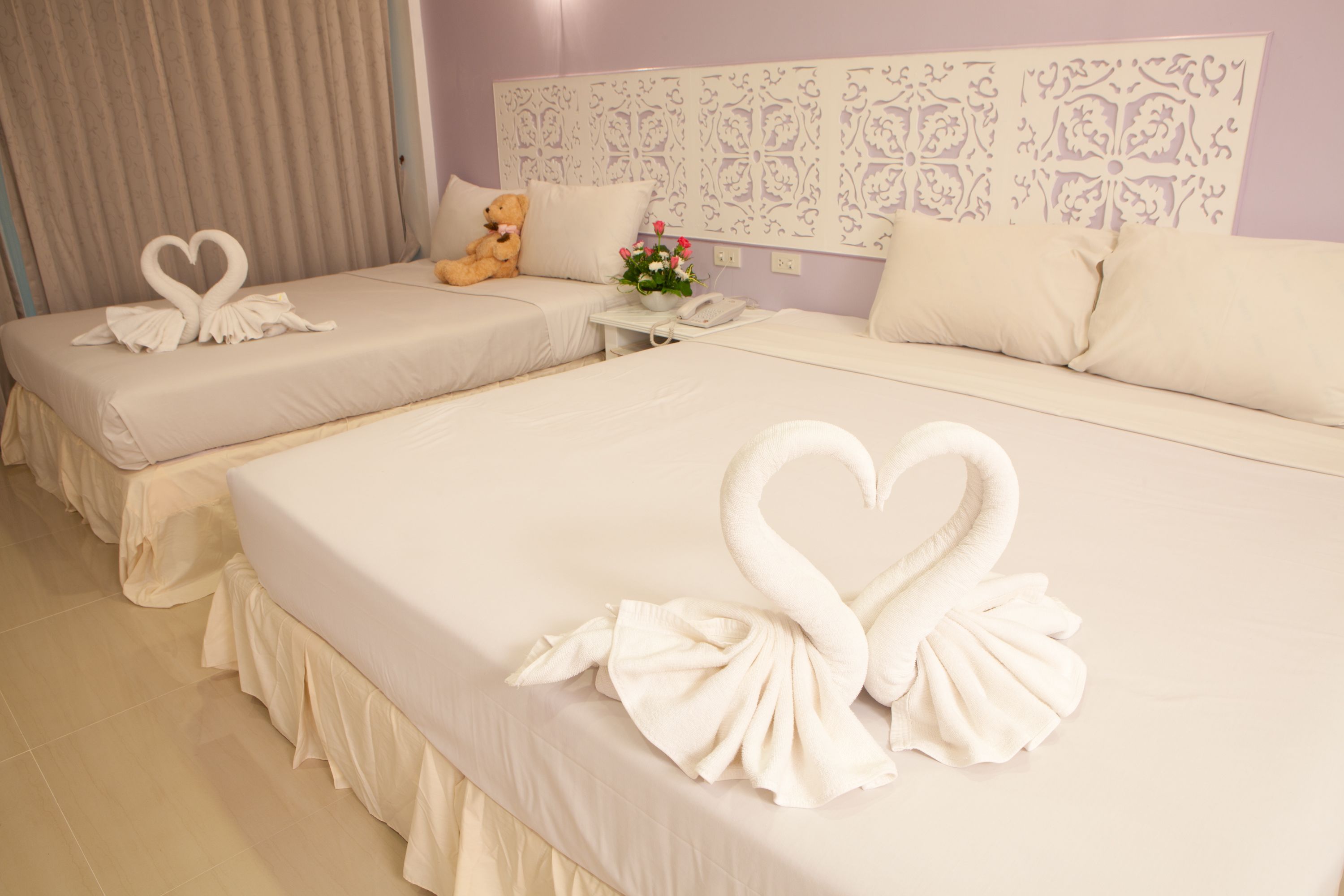 A hotel room with two beds, each with heart-shaped towels, a teddy bear, and a small table with a vase of flowers.