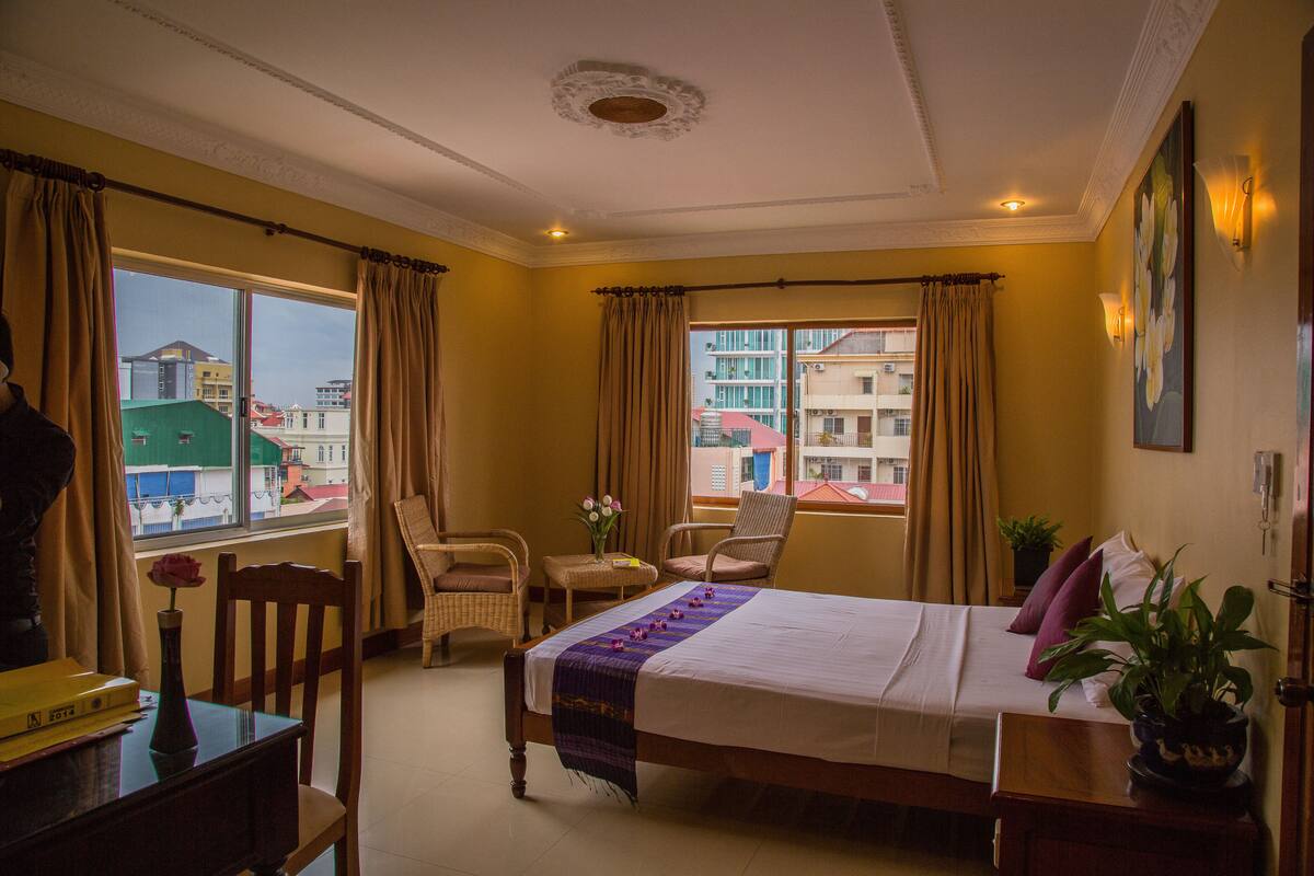 A hotel room with a bed, two chairs, a small table with a vase, and a window with curtains.