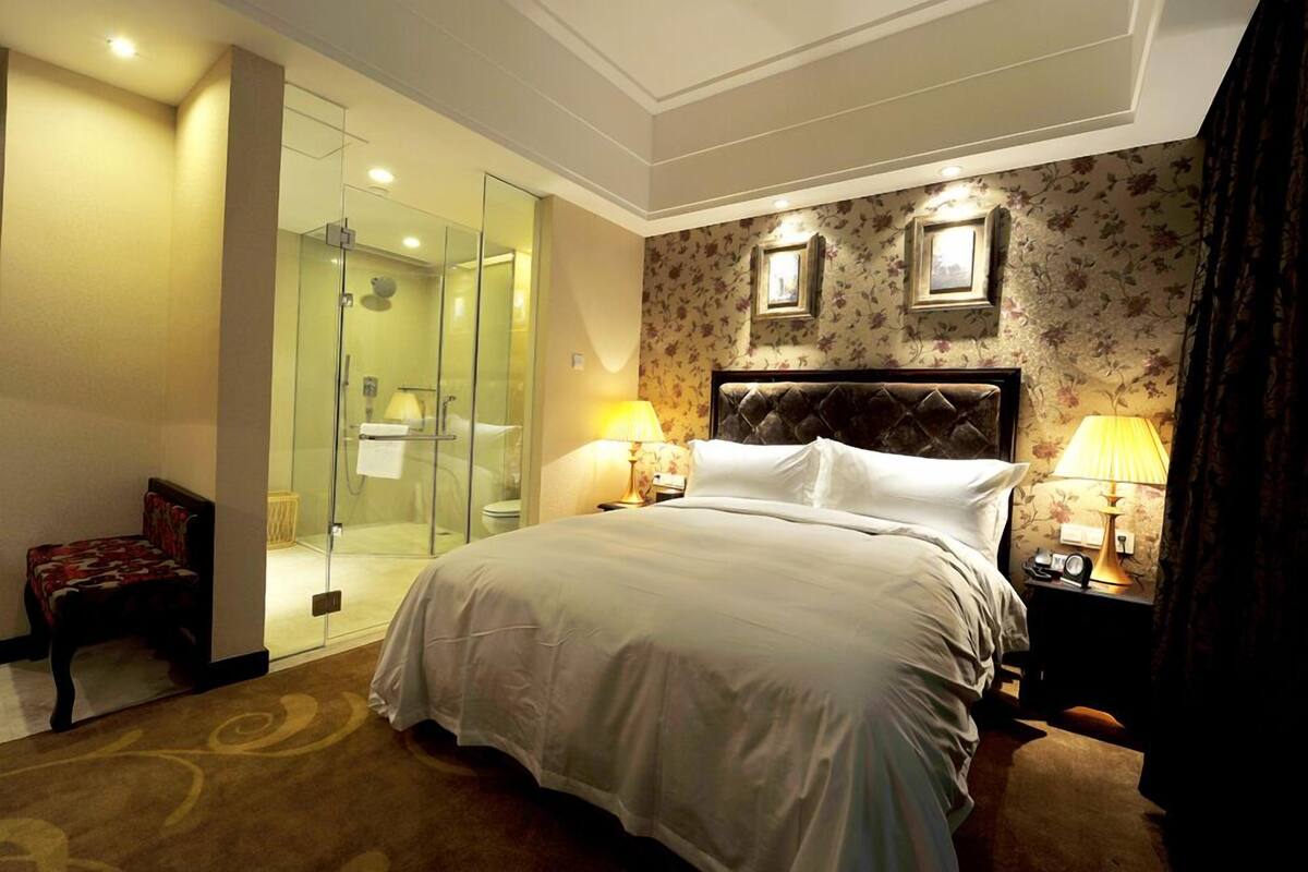 Habitació d'hotel amb un llit gran, una cabina de dutxa de vidre, una cadira i dues làmpades de nit.