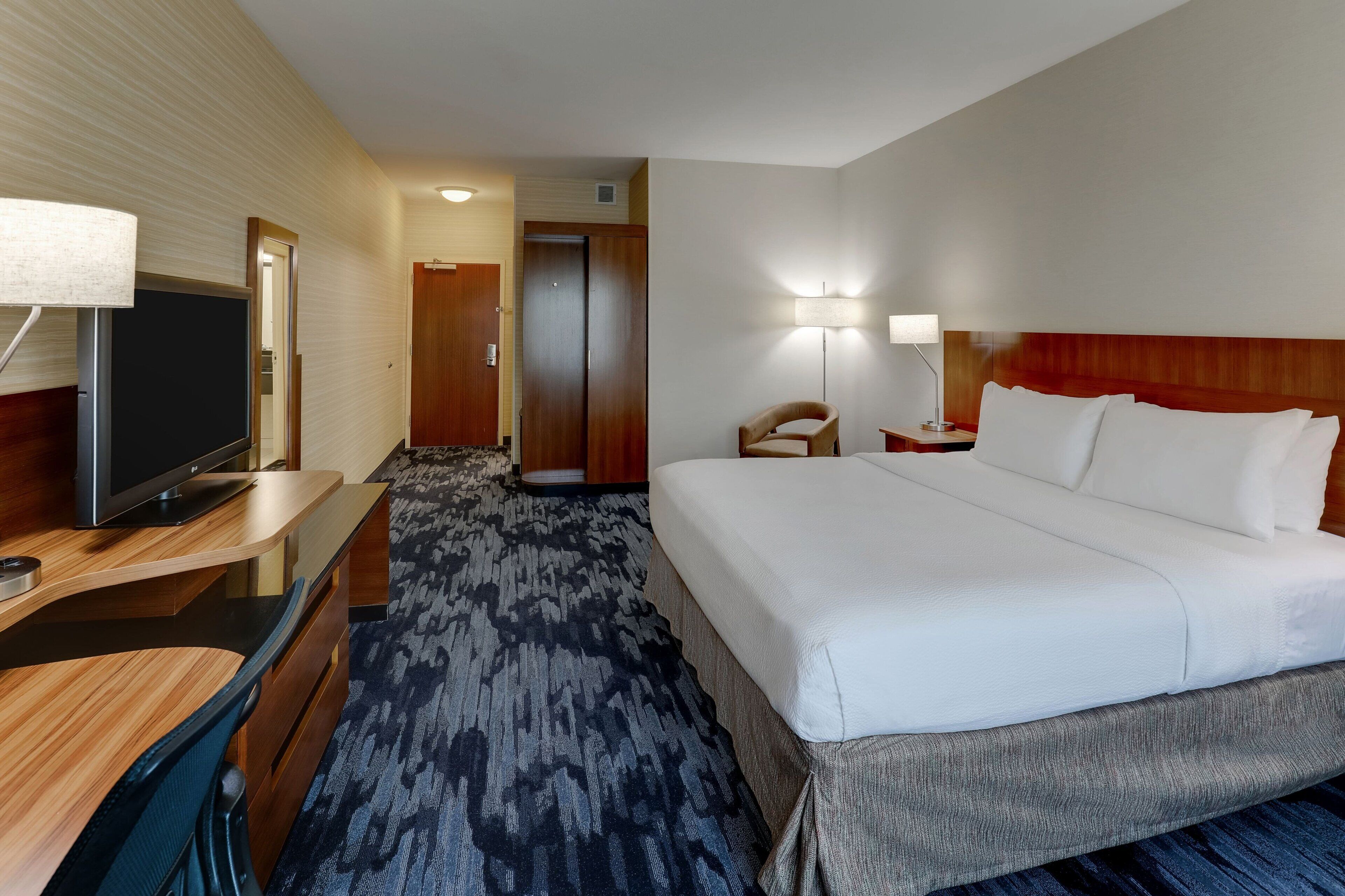 A hotel room with a large bed, a desk with a television, a chair, and a lamp.