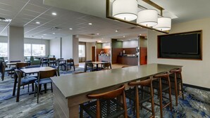 Een restaurantgedeelte van een hotel met een lange toonbank, barkrukken en tafels. Achterin is een keuken te zien en op de muur een groot scherm.