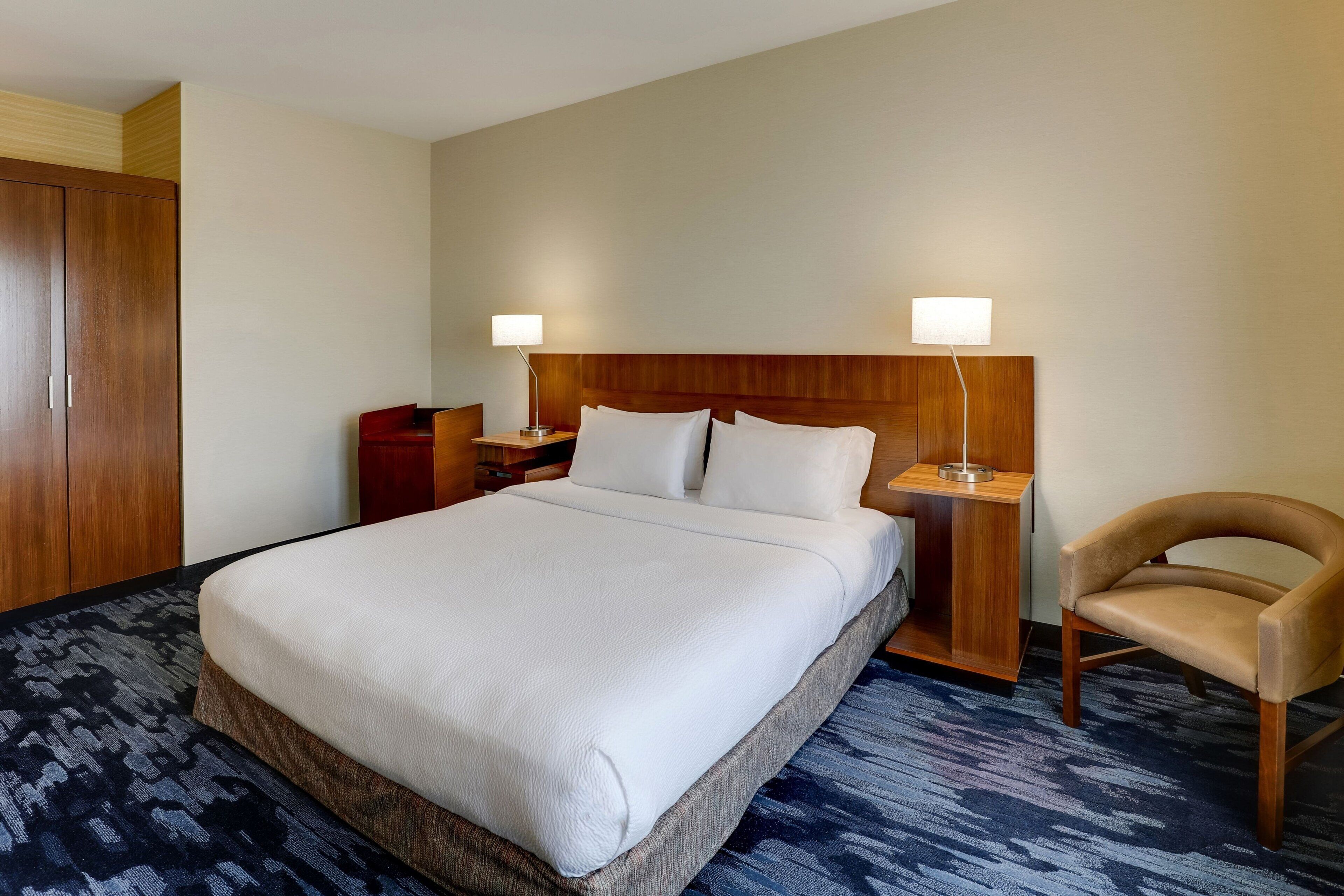 A hotel room with a large bed, two bedside tables, a wooden wardrobe, a chair, and a carpeted floor.