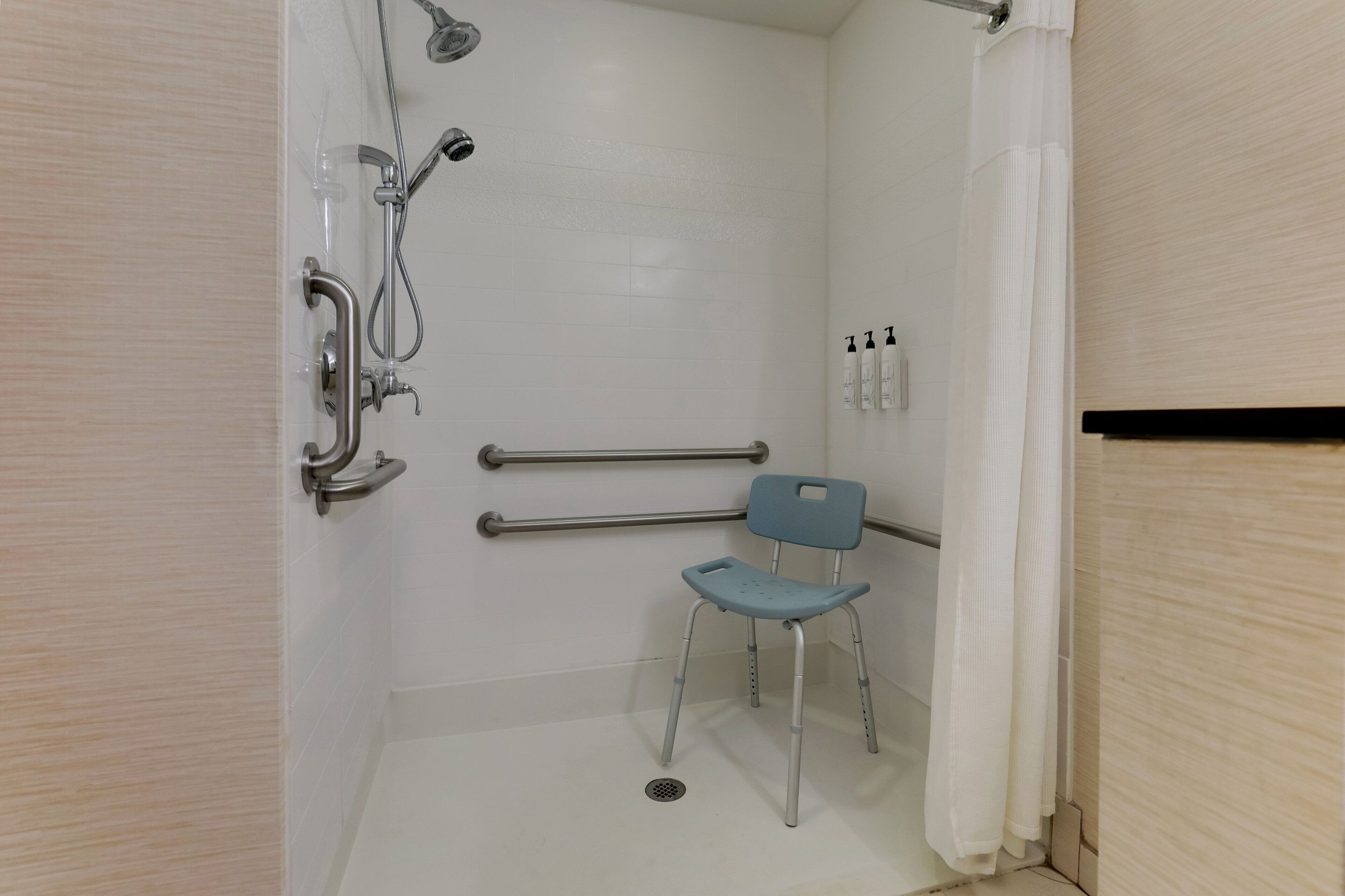 A bathroom with a shower, grab bars, a wheelchair-accessible chair, and hand sanitizer bottles.