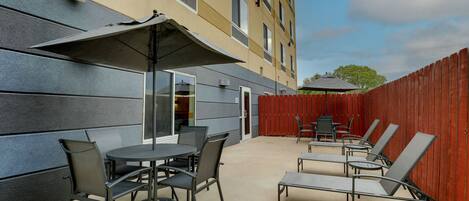 An outdoor patio area with round tables and chairs, a red wooden fence, and a building with a grey facade in the background.